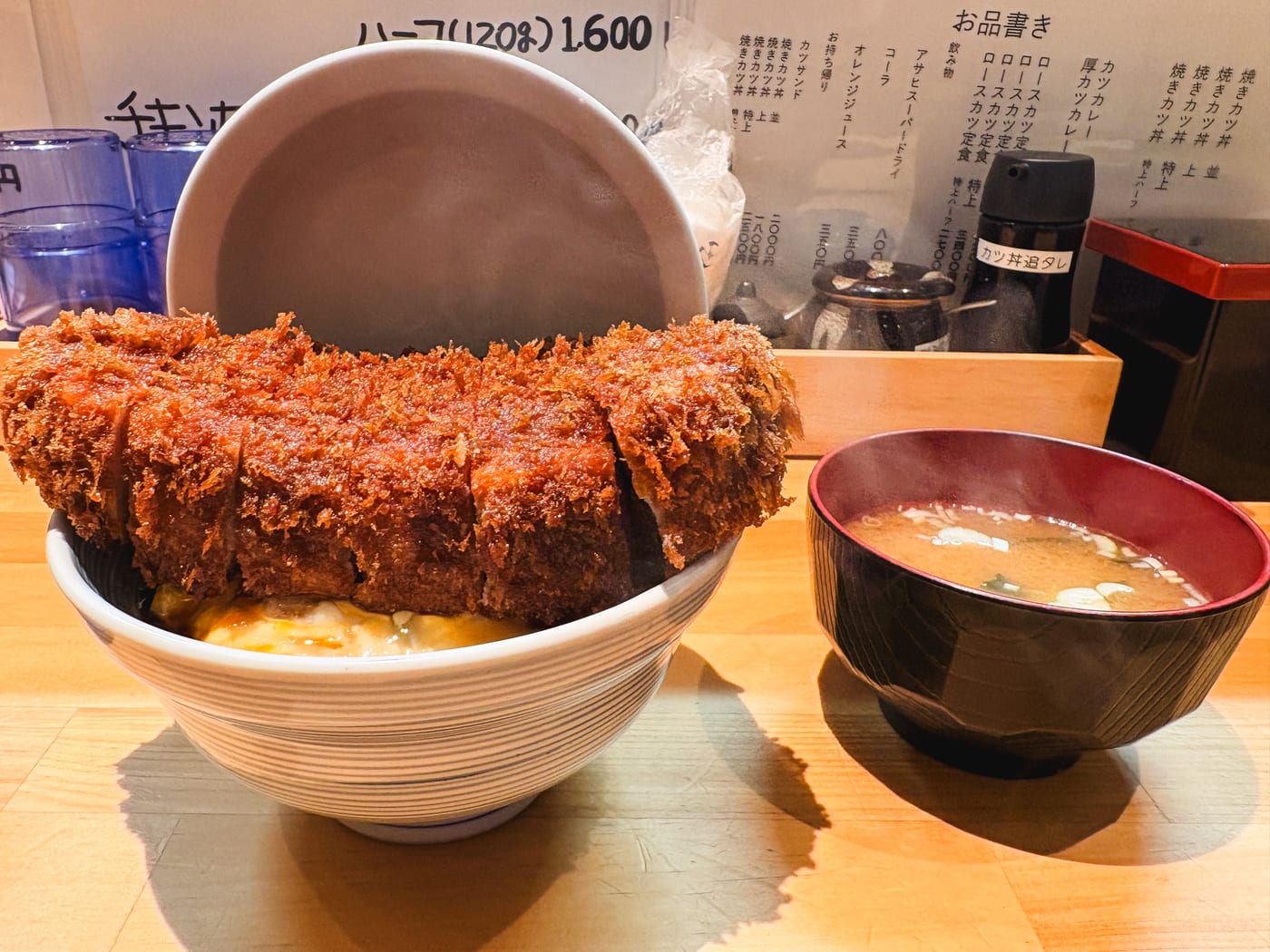 A thick pork cutlet extending well beyond the edge of its rice bowl, with miso soup alongside
