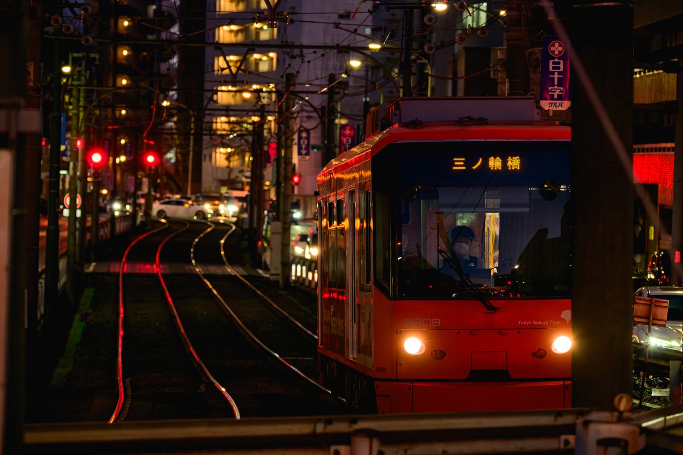 A red tram traveling through the city at night, with its lights reflected in red along the rails