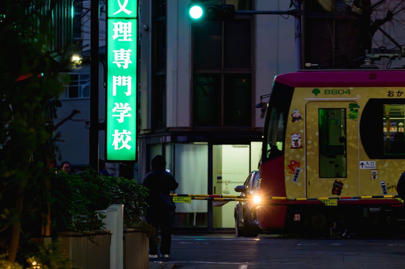 A yellow-and-burgundy tram passing in front of the green sign for Waseda-Bunri College of Arts & Sciences